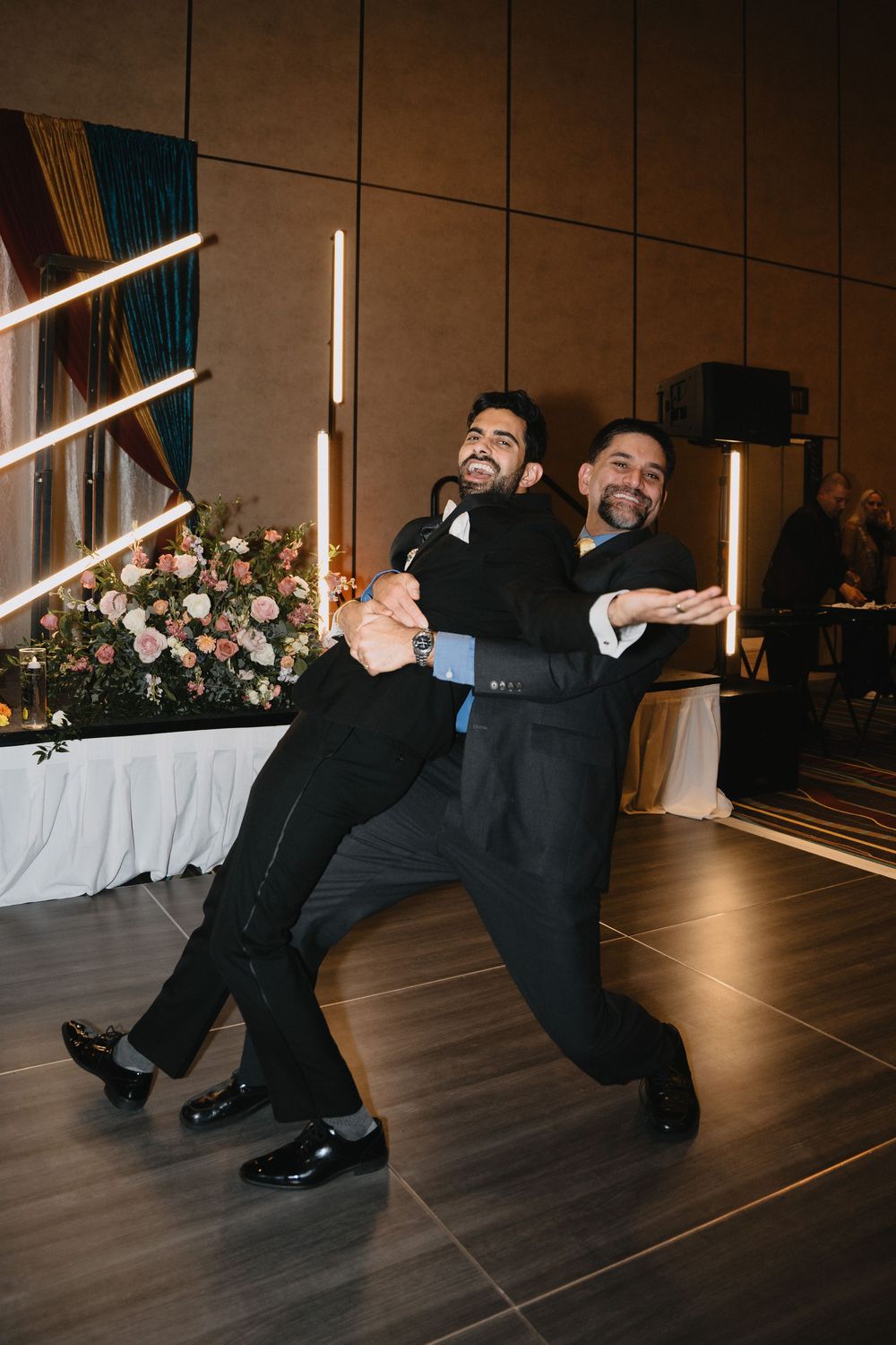Two men in black tuxedos joyfully dancing together on the wedding reception dance floor.