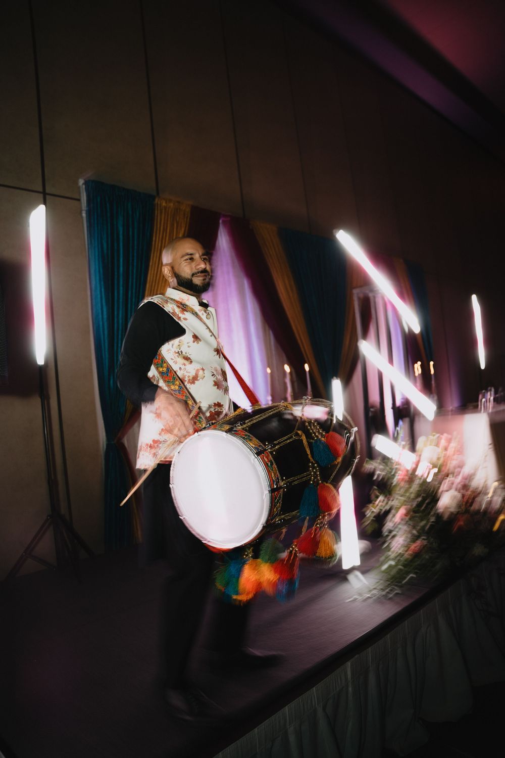A dhol drummer in traditional attire performs energetically at a wedding reception with colorful lighting.