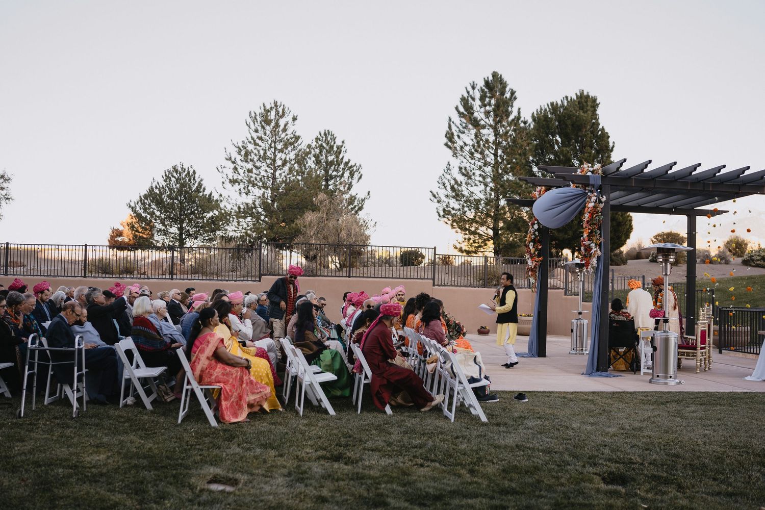 Guests in colorful traditional attire sit in white chairs at an outdoor Indian wedding ceremony at dusk.at Sandia Resort and Casino