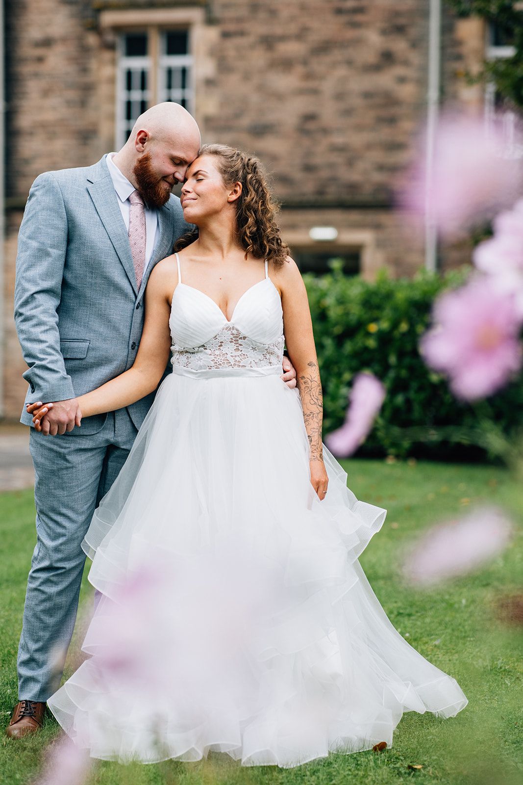 A couple shares an intimate moment in wedding attire on a lawn with pink flowers in the foreground.