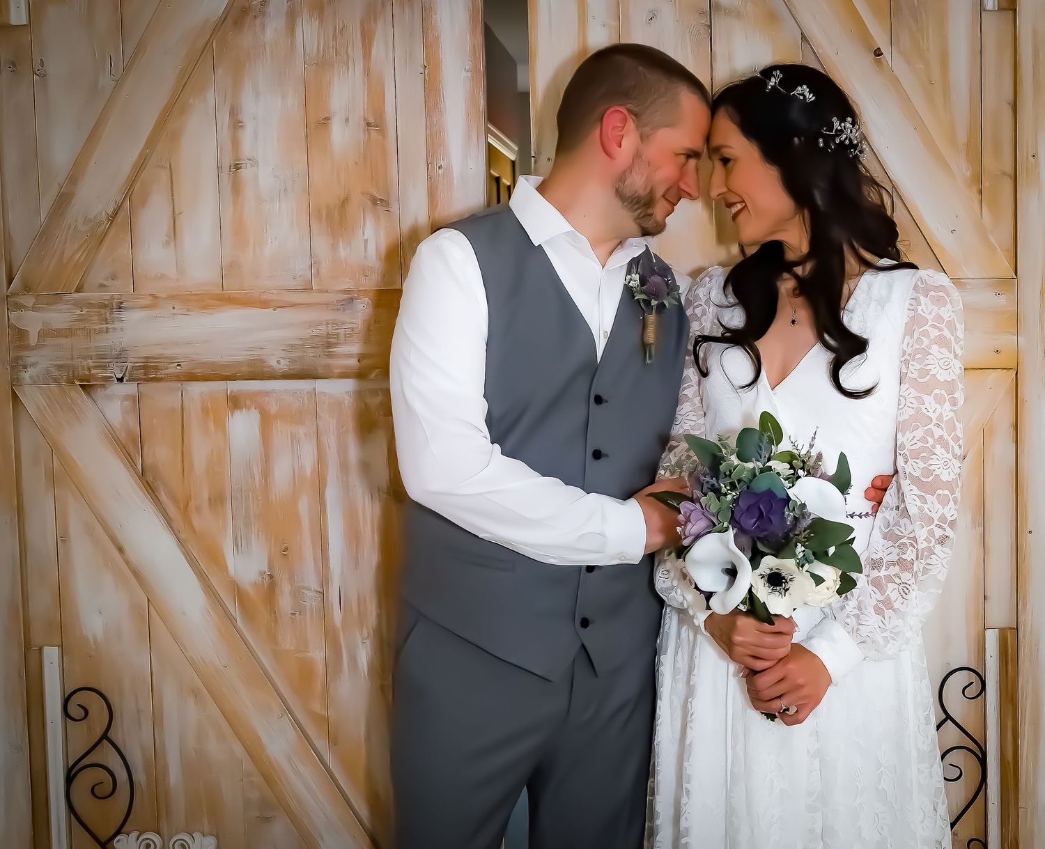 A romantic wedding couple shares an intimate moment in front of rustic barn doors with a purple and white bouquet.