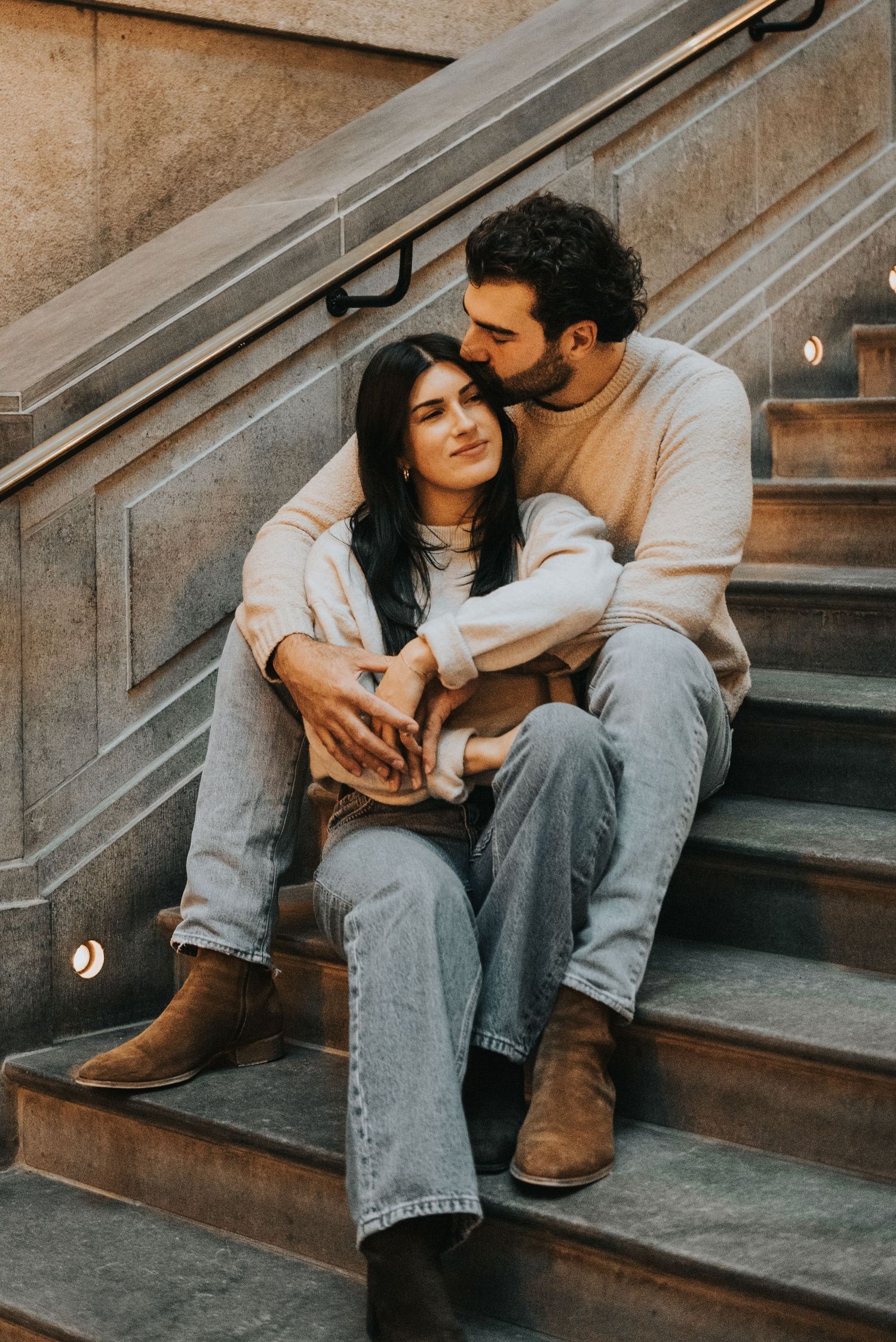 A couple sits intimately on concrete stairs in casual denim and boots with warm lighting and vintage tones.