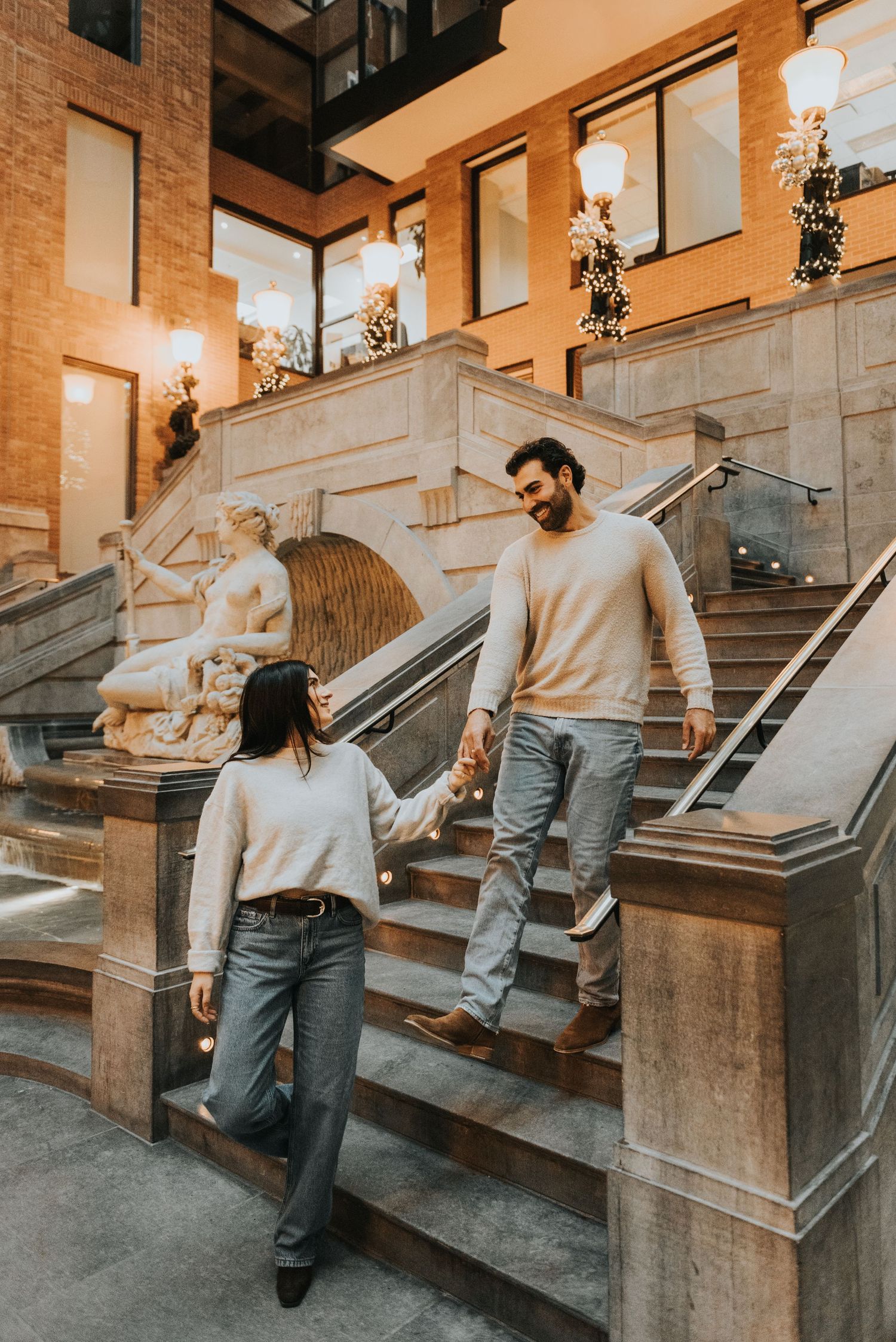 A couple in casual attire walks down stone steps outside a brick building with warm lighting.