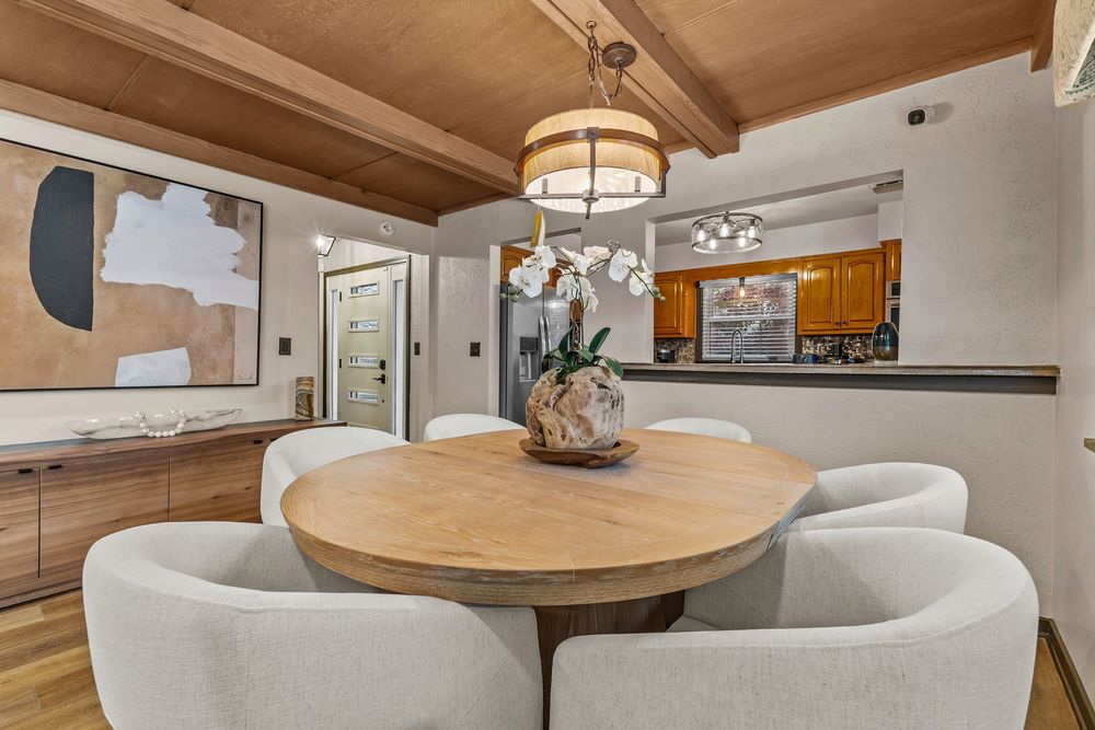 Dining area with round wooden table, white chairs, beamed ceiling, and pendant lighting.