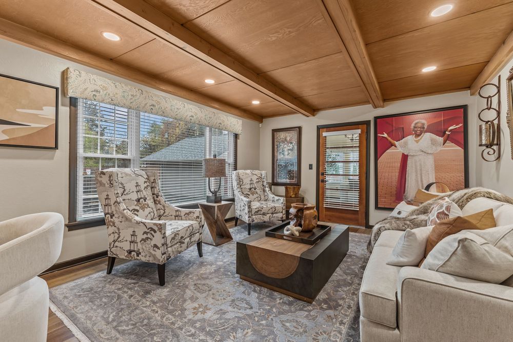 Cozy living room with beamed wood ceiling, patterned chairs, area rug, and large windows.