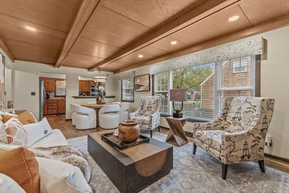 Living room with beamed ceiling, patterned armchairs, white sofa, and open kitchen view.