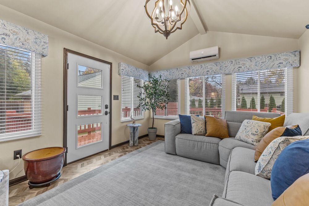 Bright sunroom with vaulted ceiling, gray sofa, floral curtains, chandelier, and mini-split AC unit.