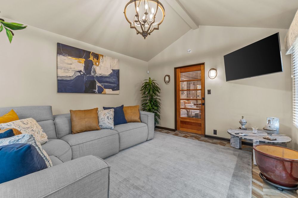 Cozy living room with vaulted ceiling, gray sofa with colorful pillows, chandelier, and wall-mounted TV.
