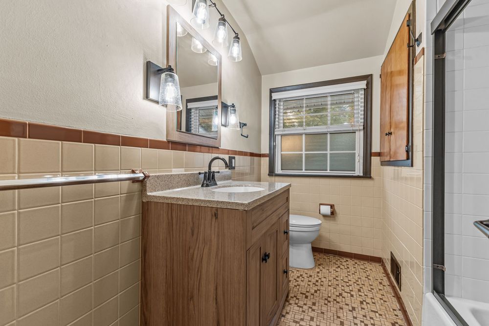 Classic bathroom with tan tile walls, wood vanity, mosaic floor tiles, and glass shower enclosure.