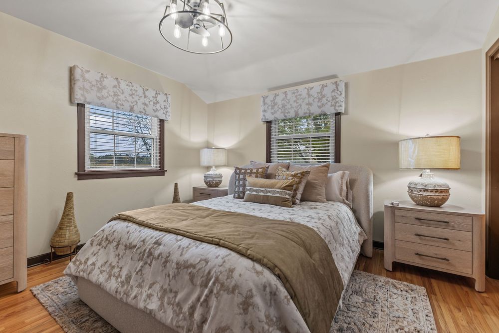 Serene bedroom with neutral tones, upholstered bed, matching nightstands, and dual windows with roman shades.