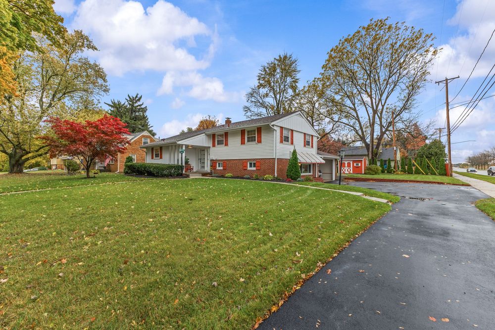 Brick split-level house with red shutters, large green lawn, mature trees, and paved driveway.