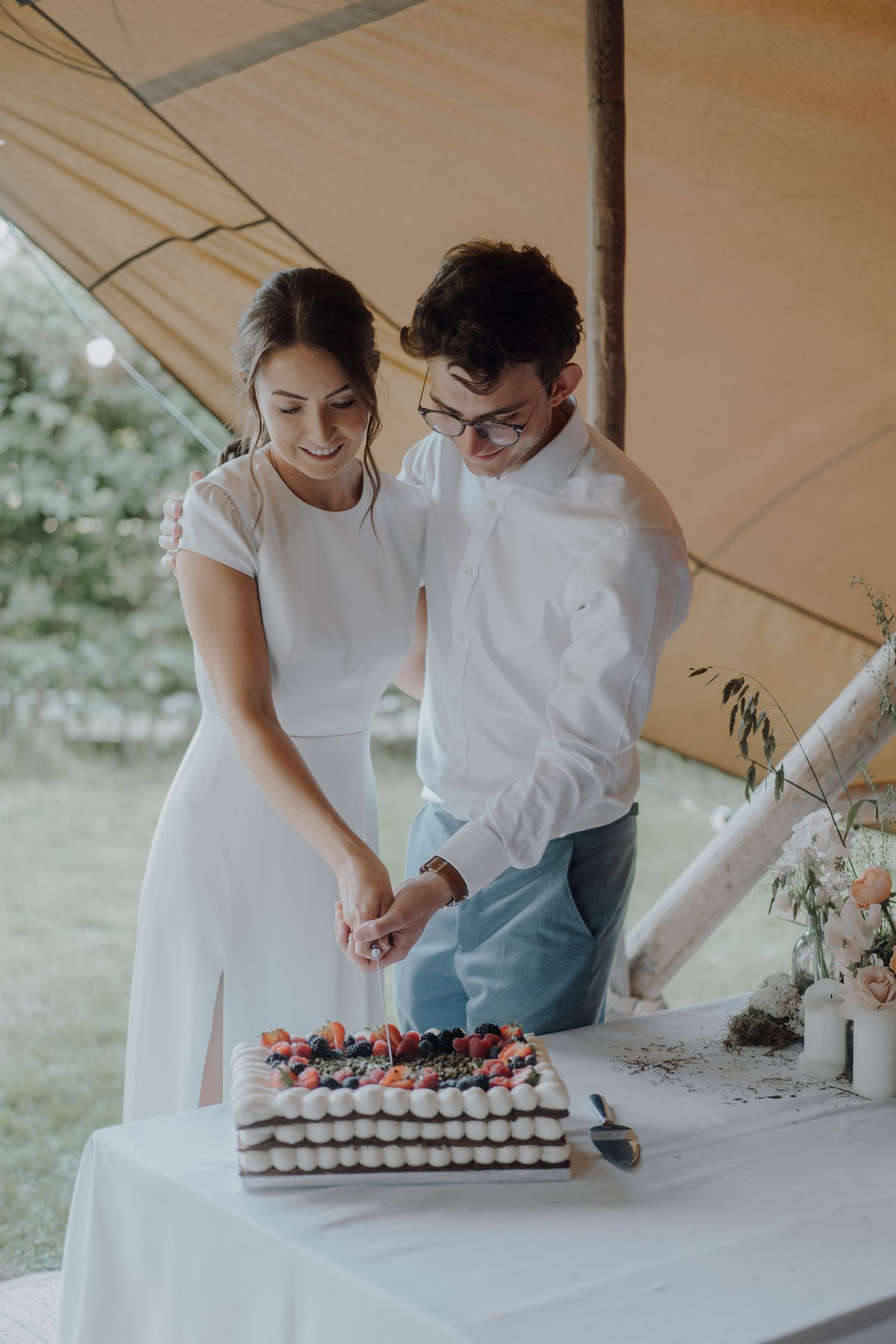 Couple in white attire cutting their decorated wedding cake under outdoor tent.