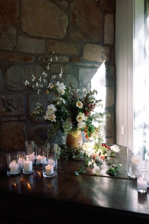 Elegant floral arrangement with white flowers and lit candles against stone wall backdrop.