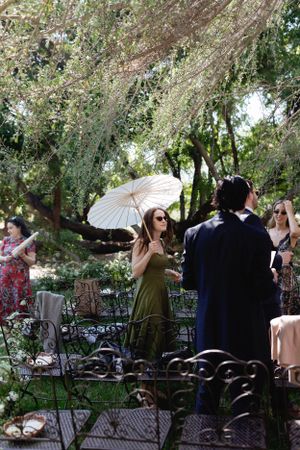 Guests mingle under flowering trees with a white parasol at an elegant garden party.