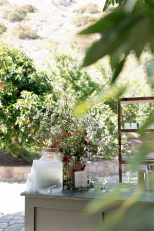 A rustic drink station with a glass beverage dispenser and floral arrangements in an outdoor garden setting.