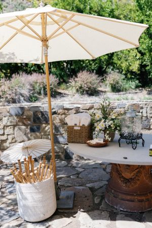Elegant outdoor patio setup with white umbrella, parasols in basket, and stone wall details in garden setting.