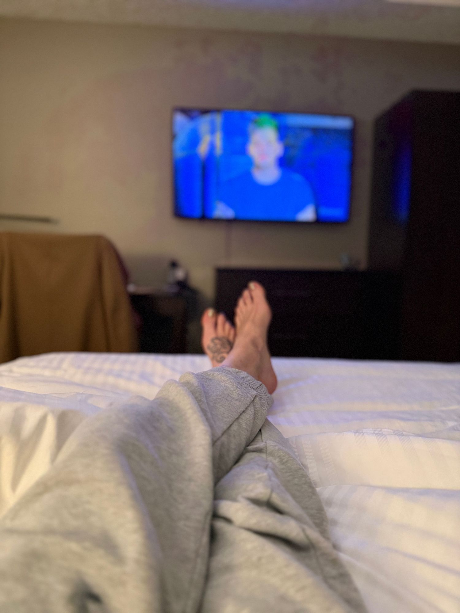 Person relaxing on white bedding while watching TV with blue screen glow visible in dark room.