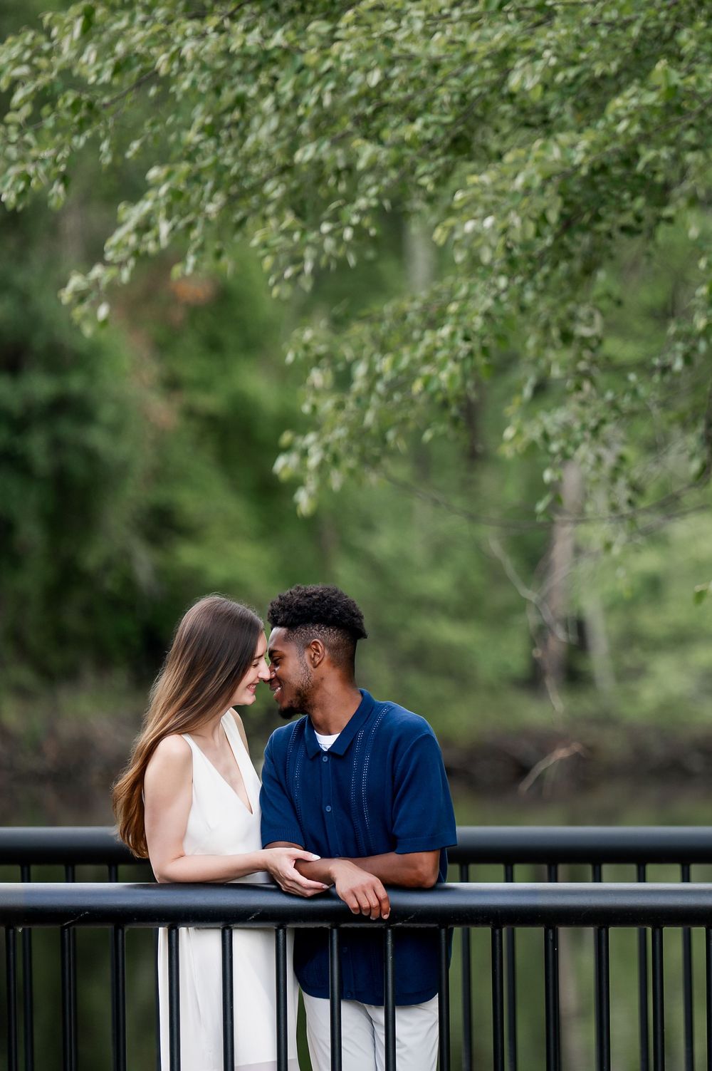 Romantic couple sharing tender moments along a railing in a lush green park setting.