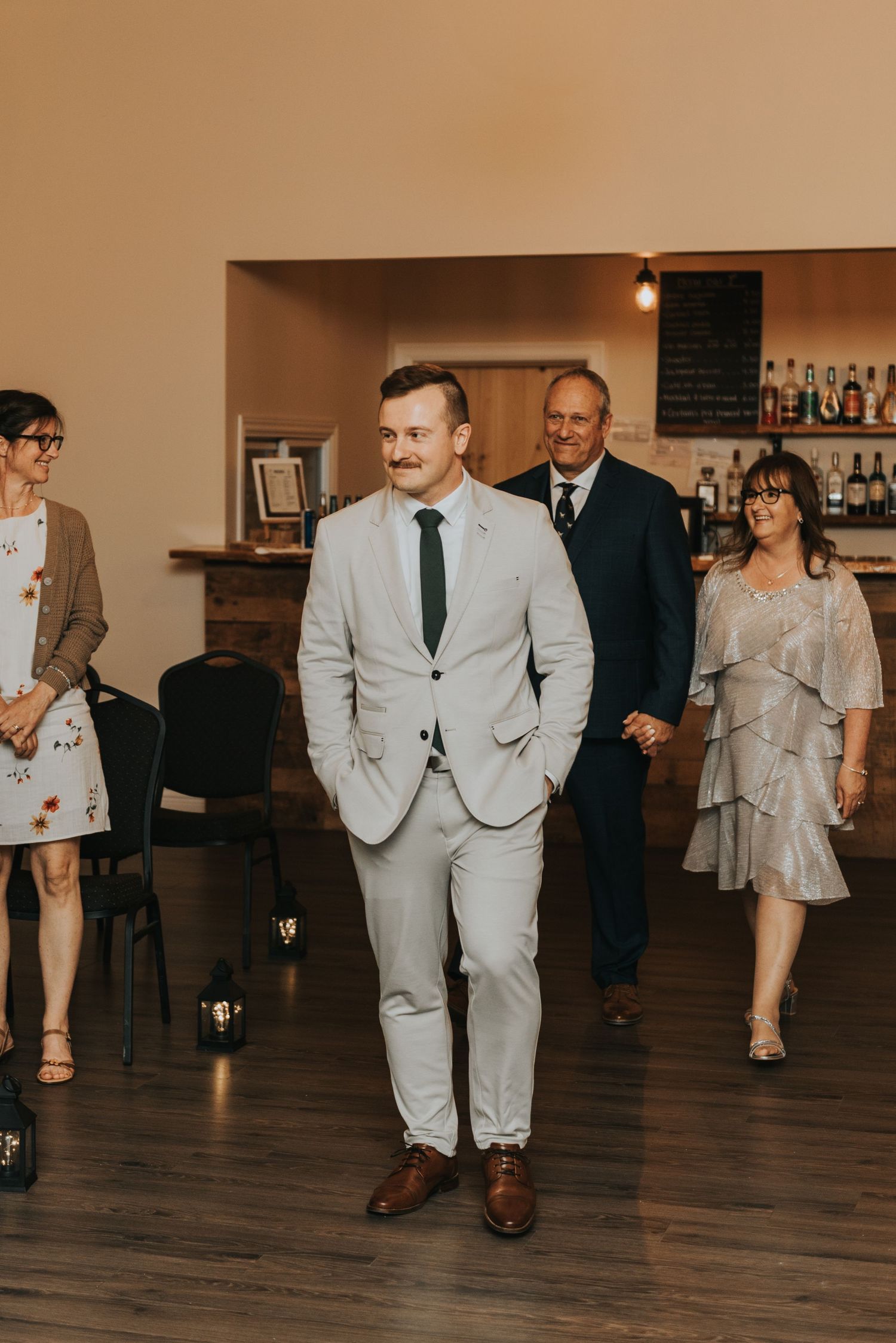 Wedding guests in light gray suits and formal attire walk across a wooden floor at an indoor reception venue.