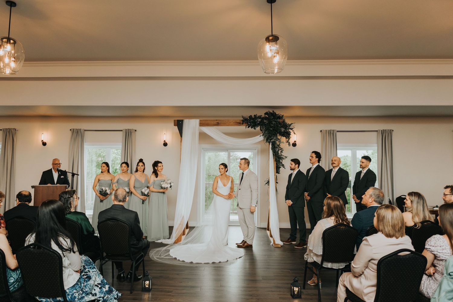 Indoor wedding ceremony with wedding party at altar and guests seated in white chairs under crystal pendant lights.