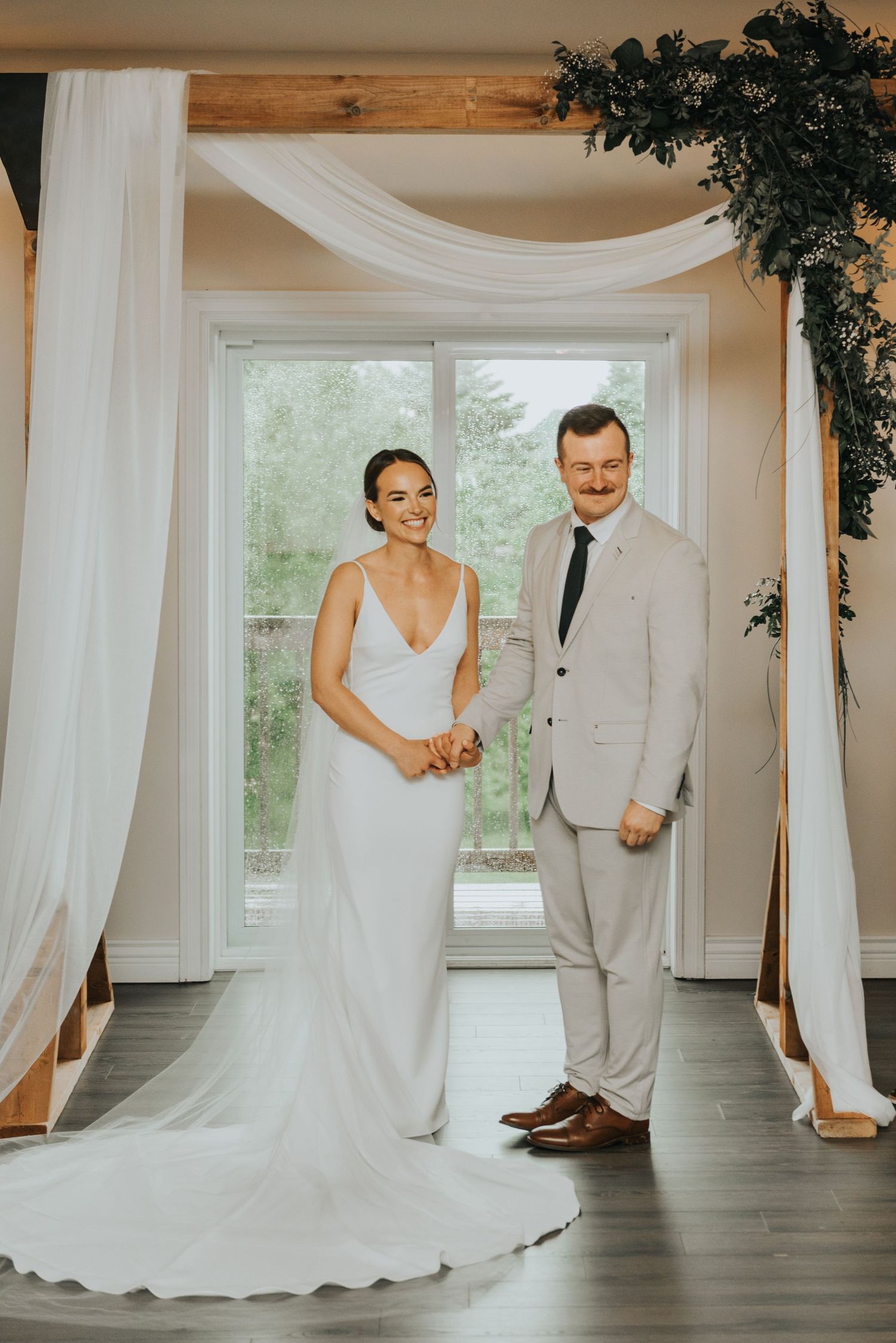 A wedding couple stands together in an elegant white room with flowing drapery and natural light from large windows.