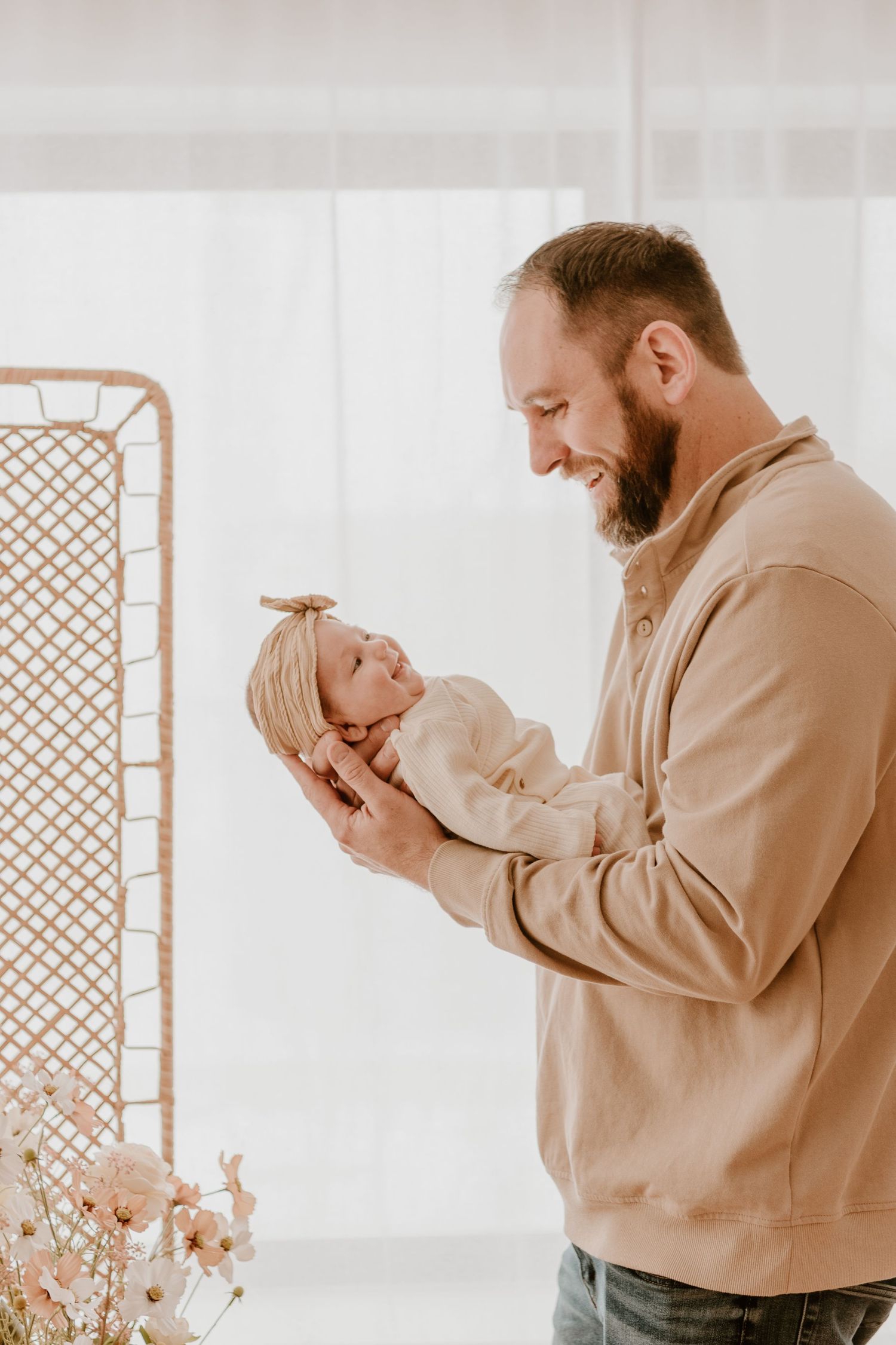 Person in tan sweatshirt holding small baby while standing next to rattan furniture.