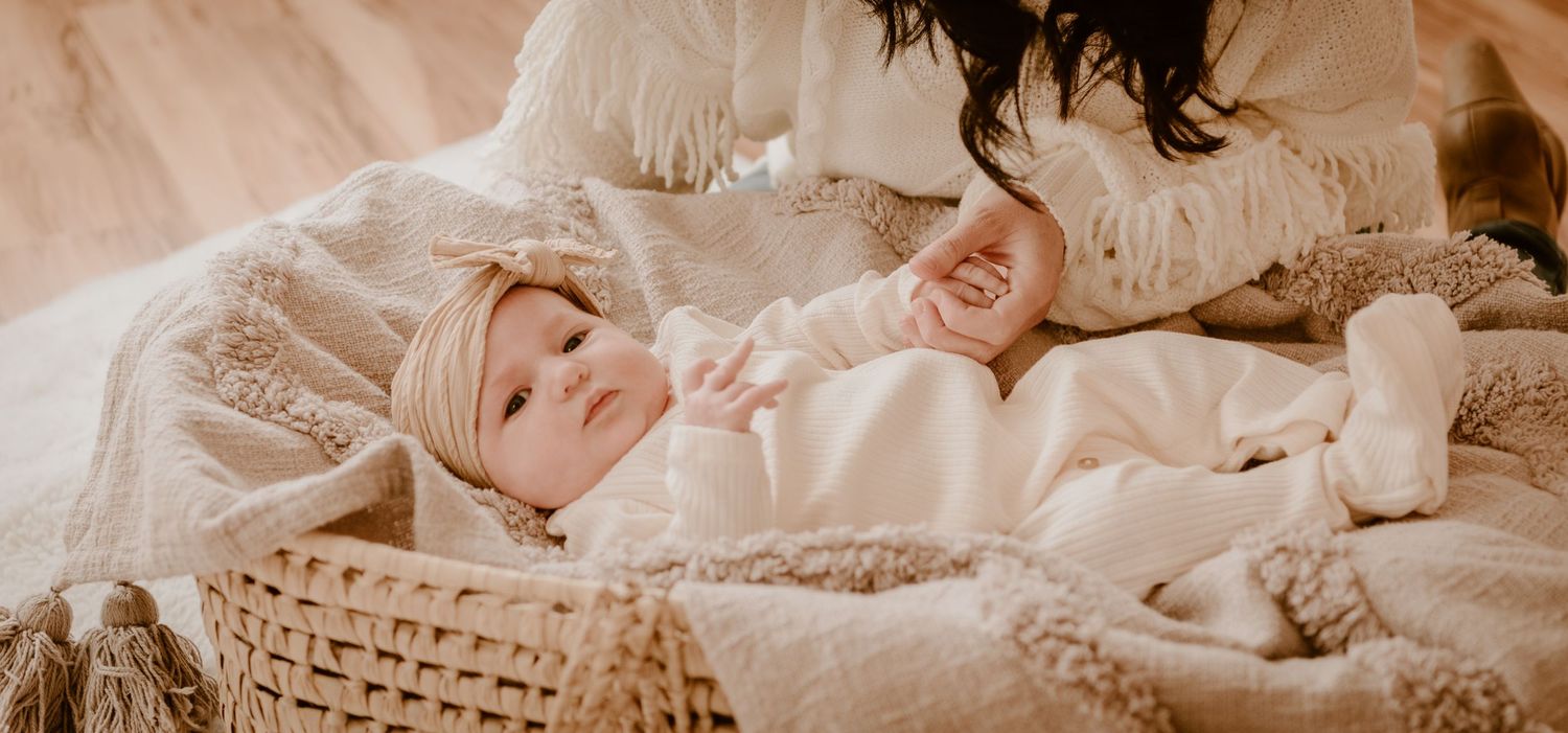 Baby lying peacefully in woven basket surrounded by soft neutral textiles and blankets.