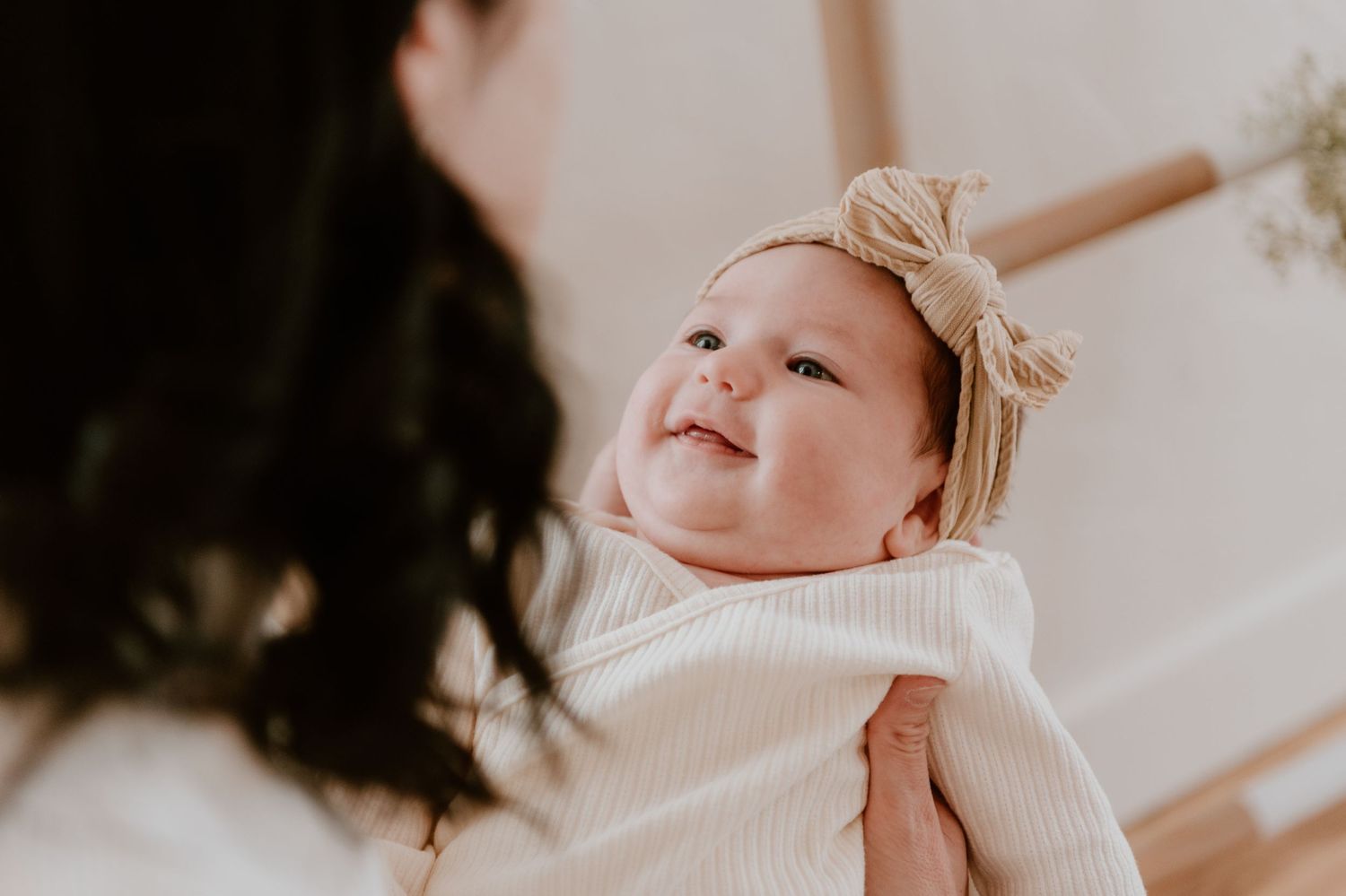 Baby wearing white sweater and bow smiles while being held up against white backdrop.