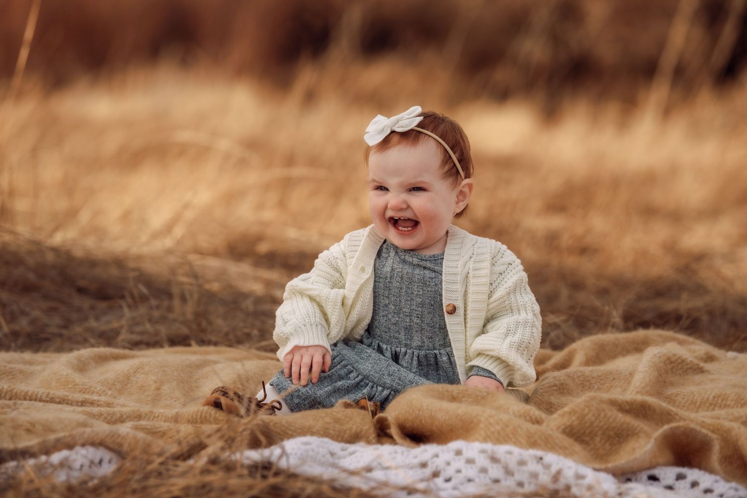 A baby wearing a gray dress sits and laughs joyfully in a golden wheat field on a sunny day.