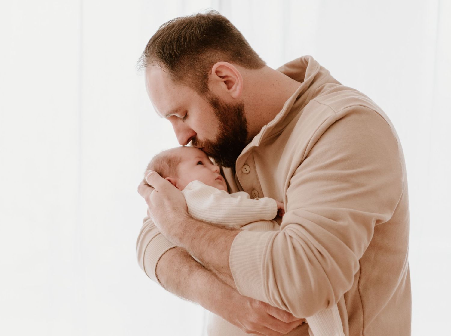 Person in beige sweatshirt holds infant close in tender embrace against white background.
