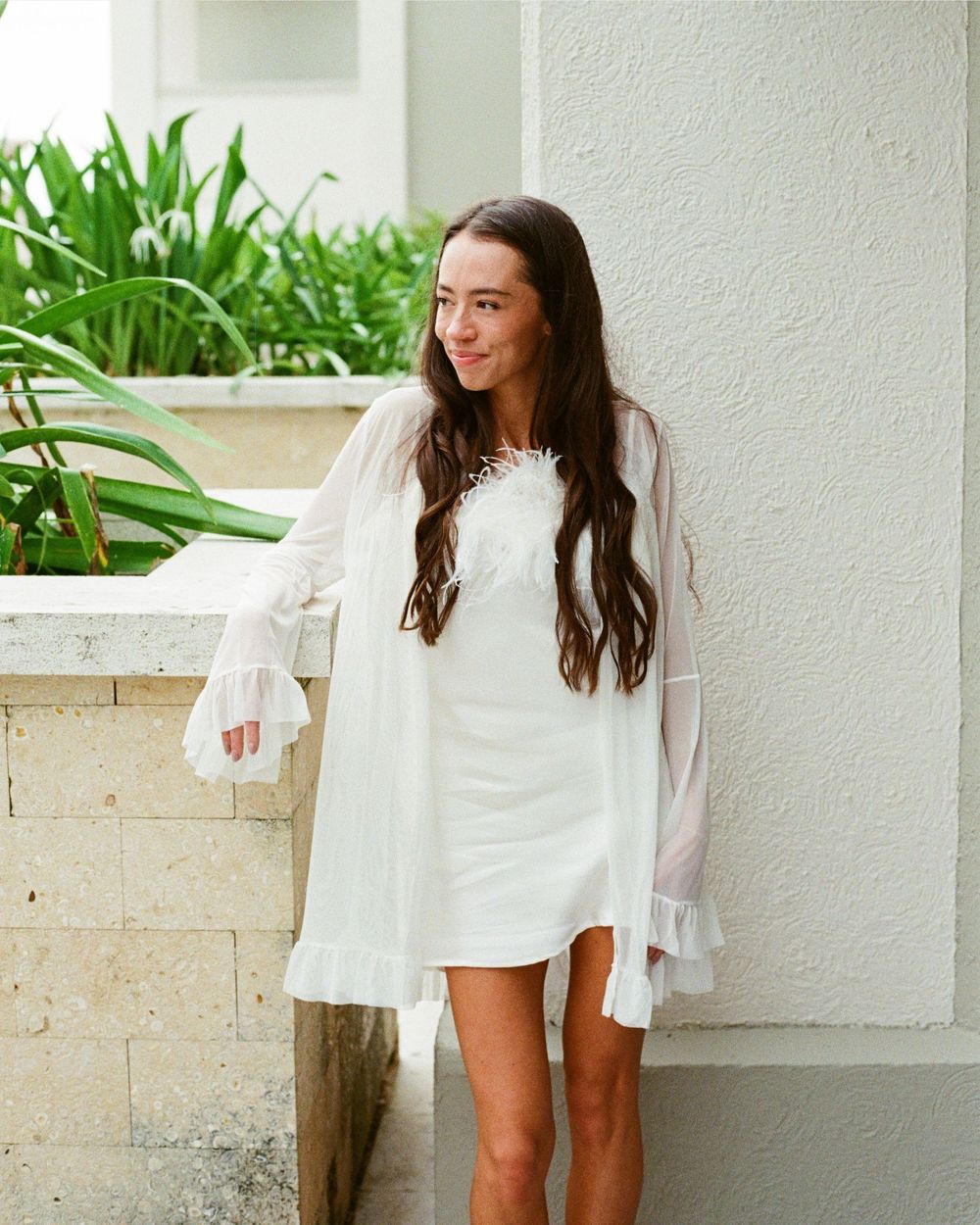 A woman in a white dress and robe stands against a wall with tropical plants.