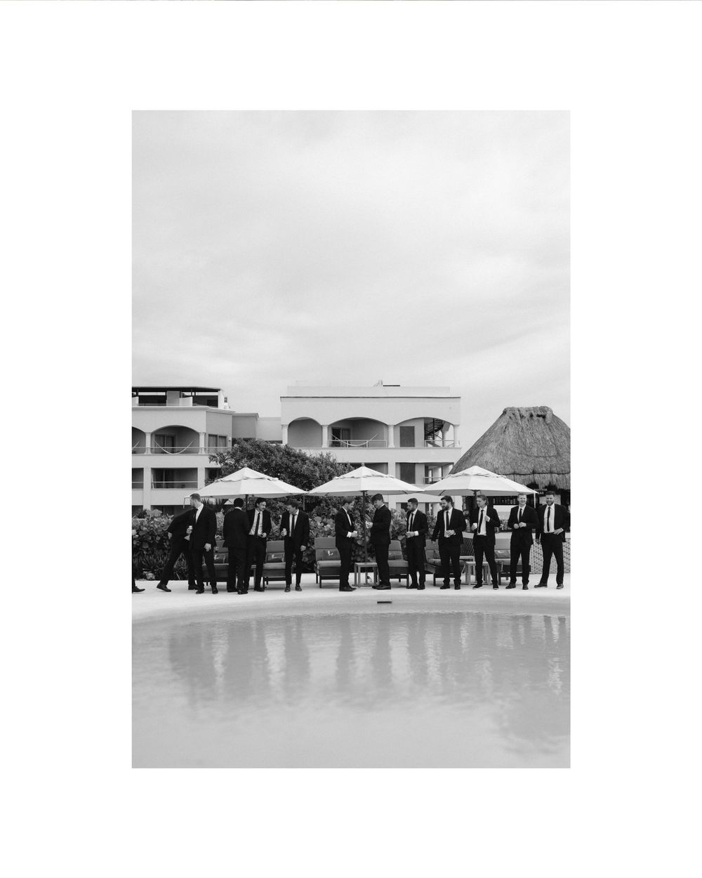 Black and white photo of a poolside wedding reception with guests under umbrellas.