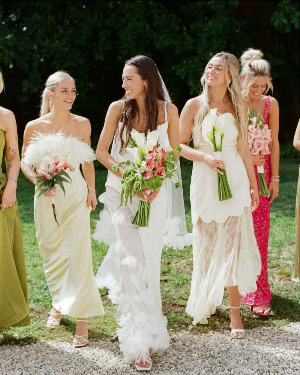 Wedding party members in light-colored dresses walking together while holding bouquets.