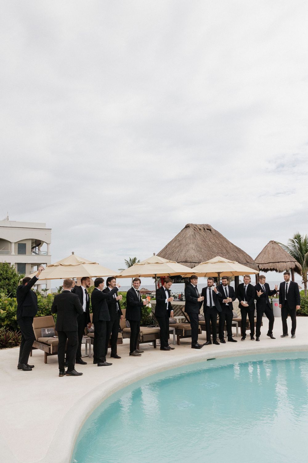 Wedding party in formal black attire lined up along a tropical beachfront pool at sunset.