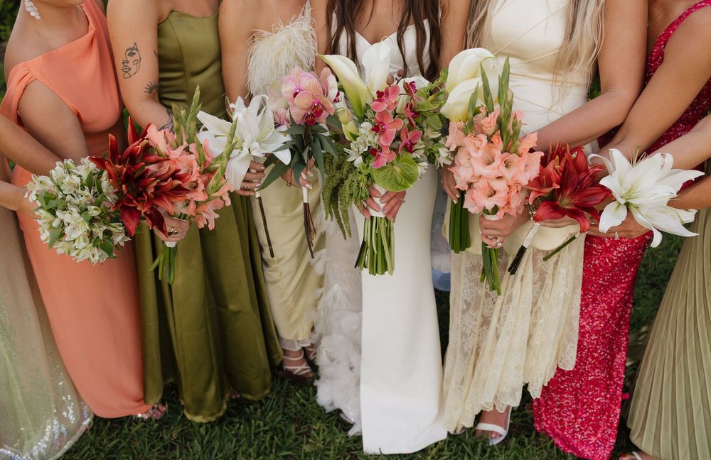 Wedding party members hold colorful bouquets featuring pink and red flowers against white dresses and green attire.