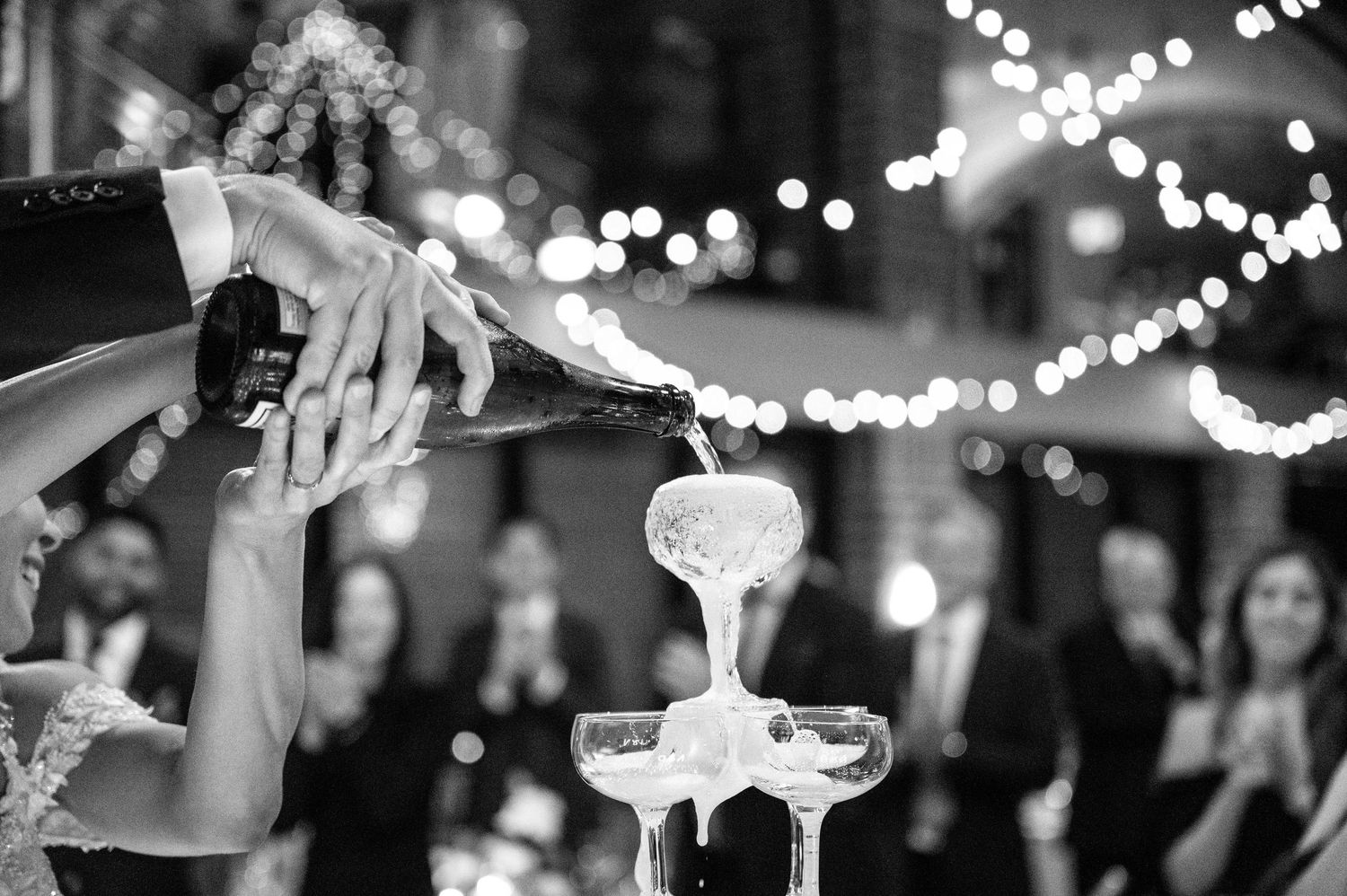 Champagne being poured into tower of glasses at elegant wedding reception with bokeh lights.
