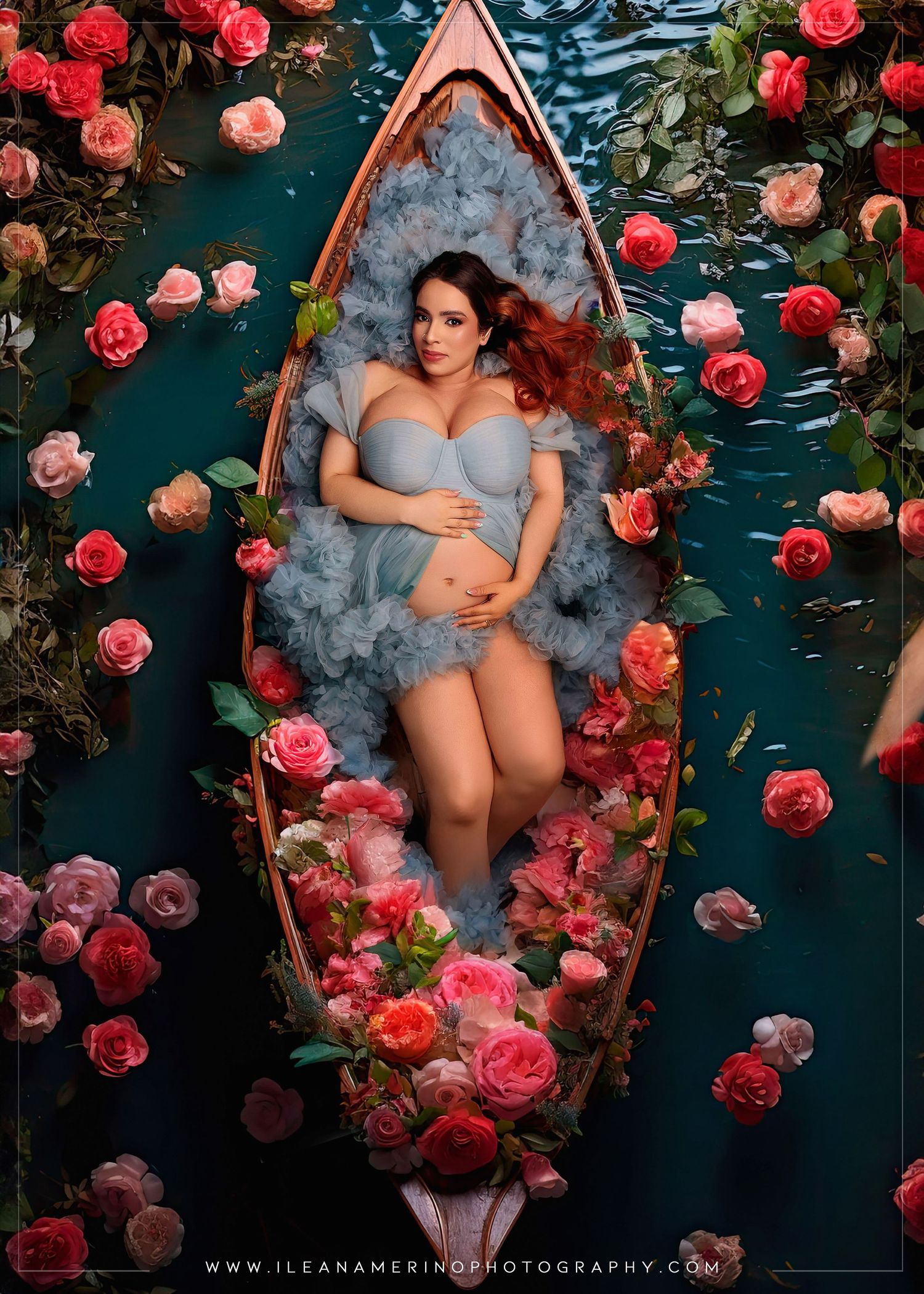 Pregnant woman in blue lingerie reclines in a flower-filled wooden boat surrounded by pink roses on teal water.