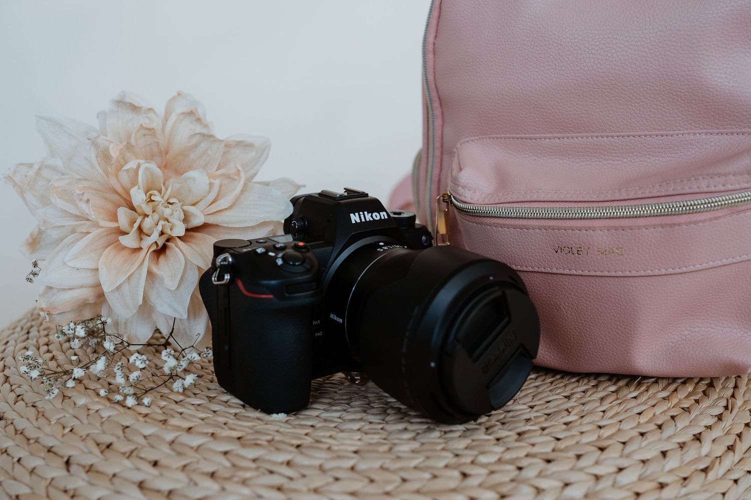 Black Nikon camera with lens sits next to pink leather backpack and peach flower on woven surface.
