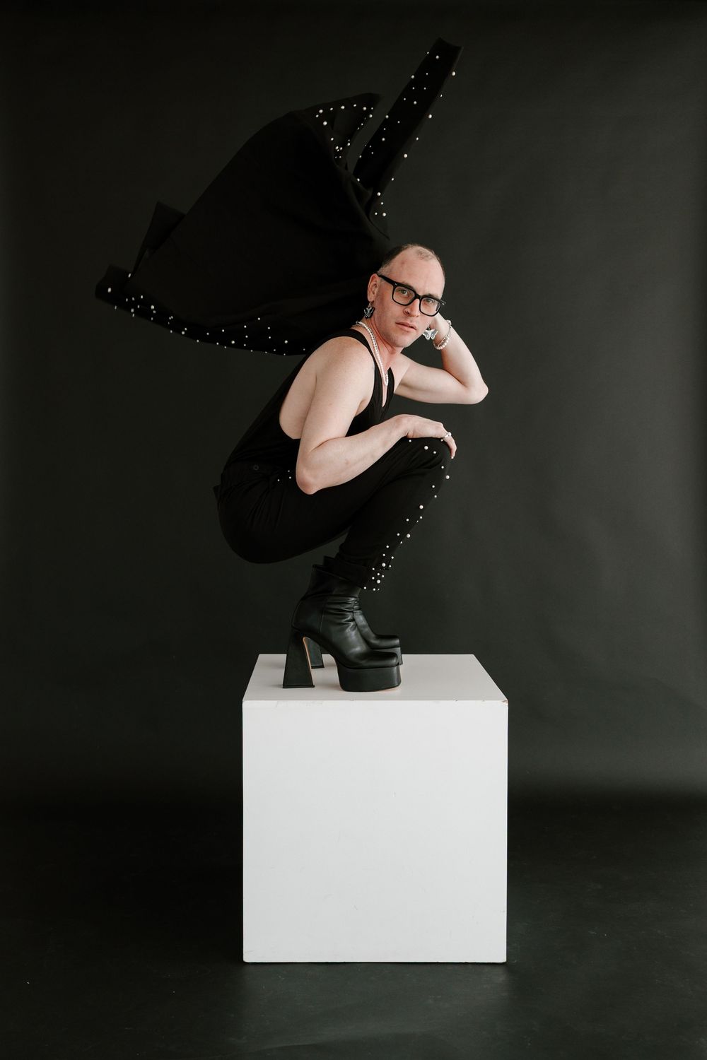 Figure in black outfit and platform boots poses artistically on white cube pedestal against dark background.
