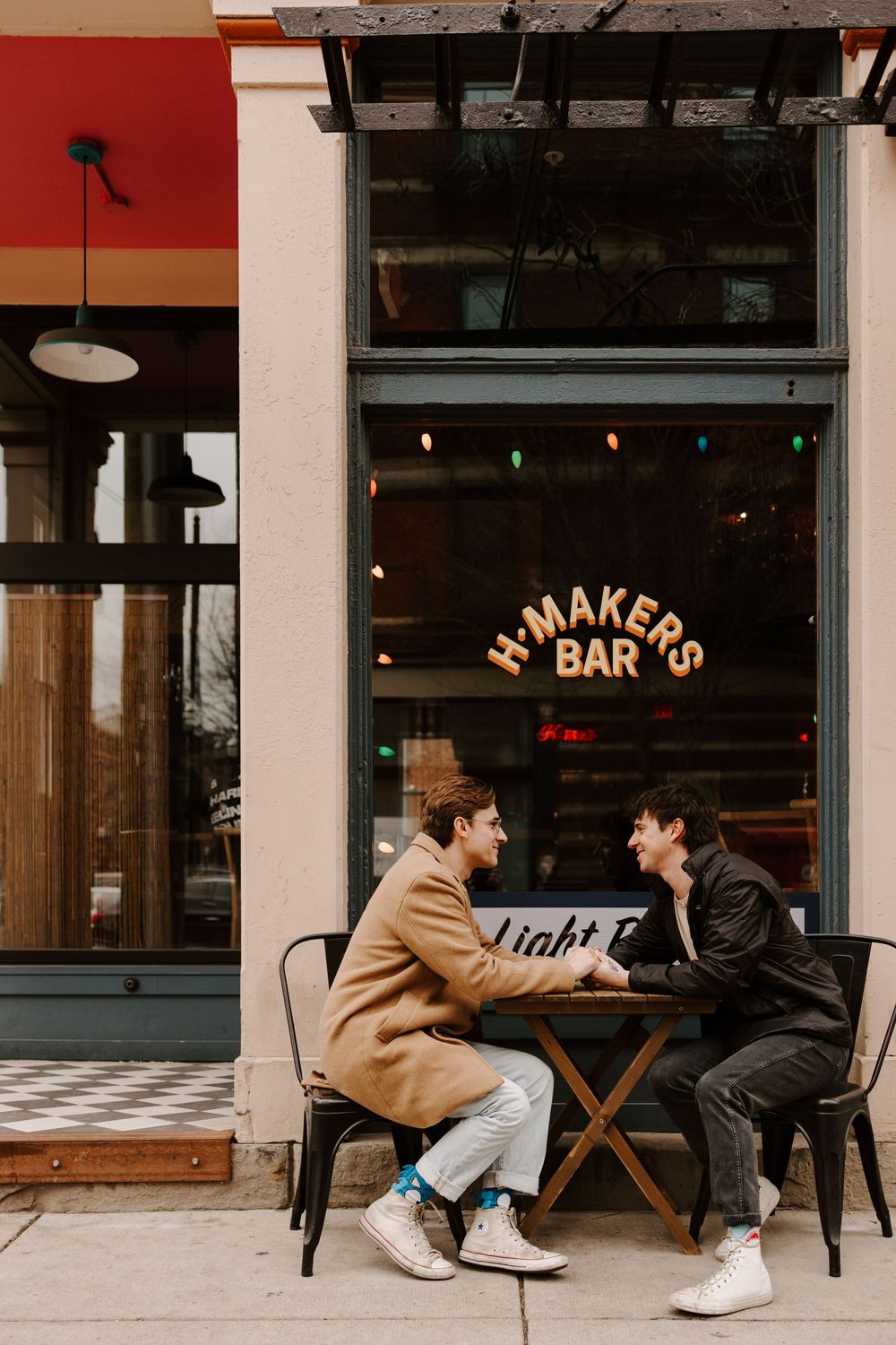 Two people sit at a cafe table outside a bar with a vintage storefront sign in warm evening lighting.