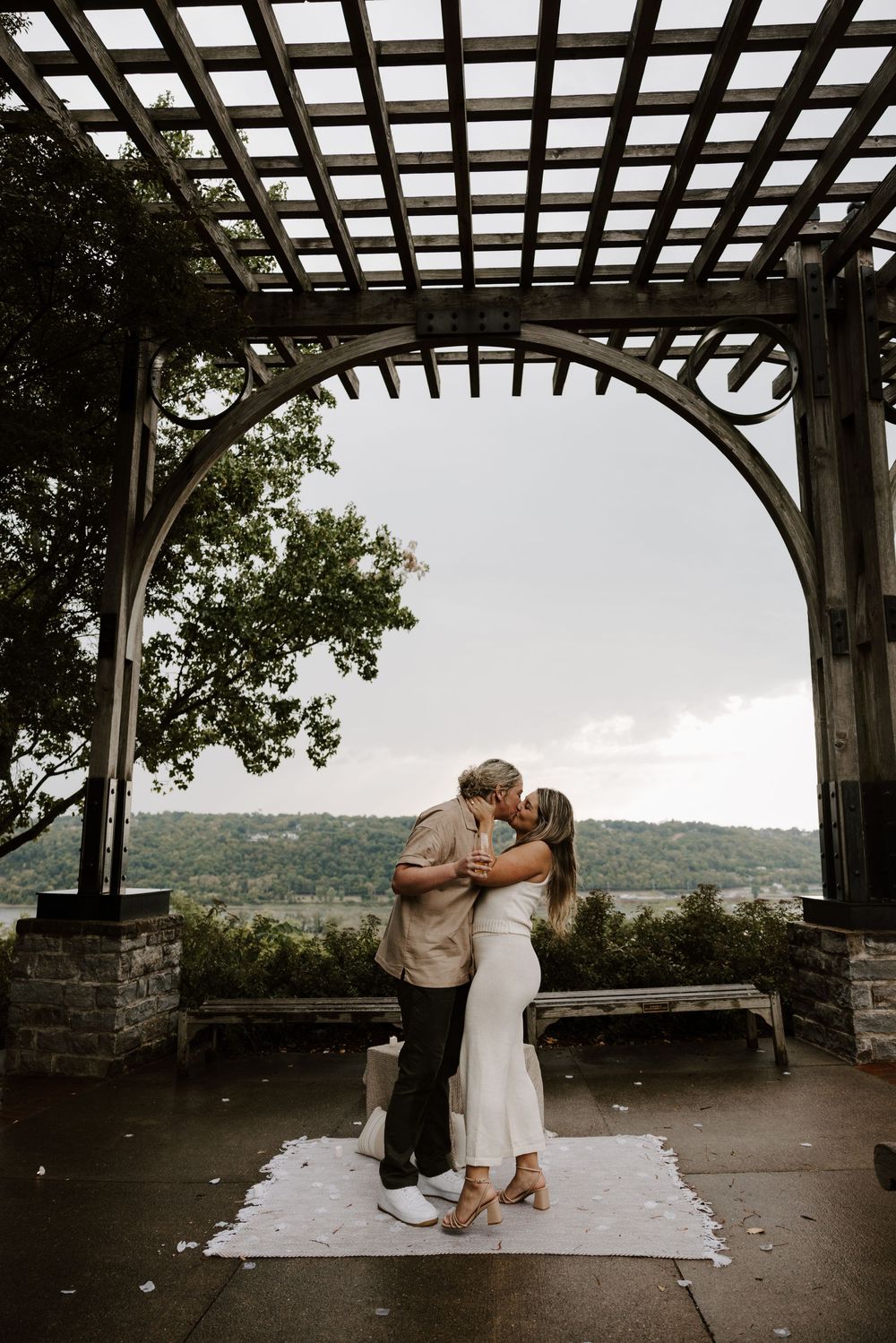 A romantic couple shares a tender moment under a wooden pergola overlooking a scenic mountain landscape at sunset.