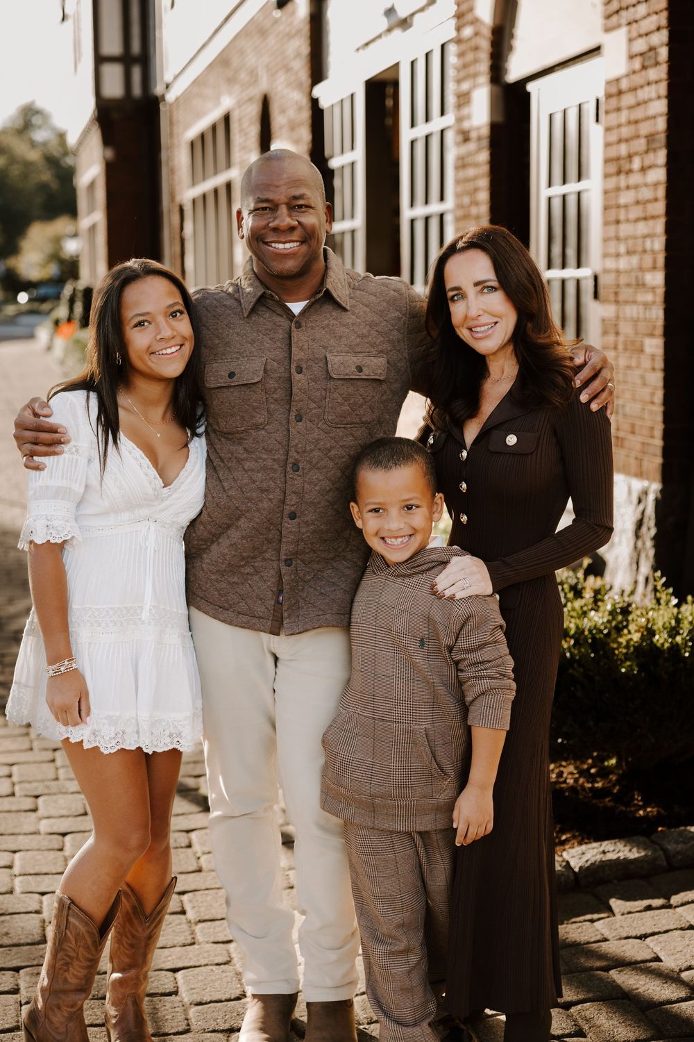 Family wearing coordinated brown and white outfits poses together against brick wall backdrop.