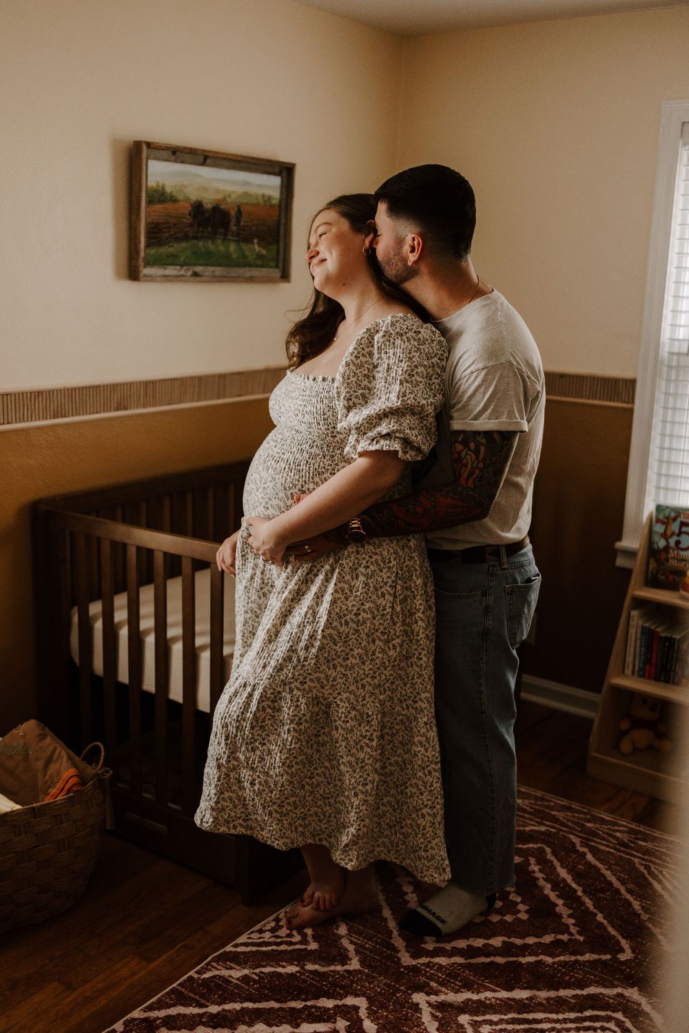 A loving couple shares an intimate moment in a nursery room with a dark wood crib and landscape painting on the wall.