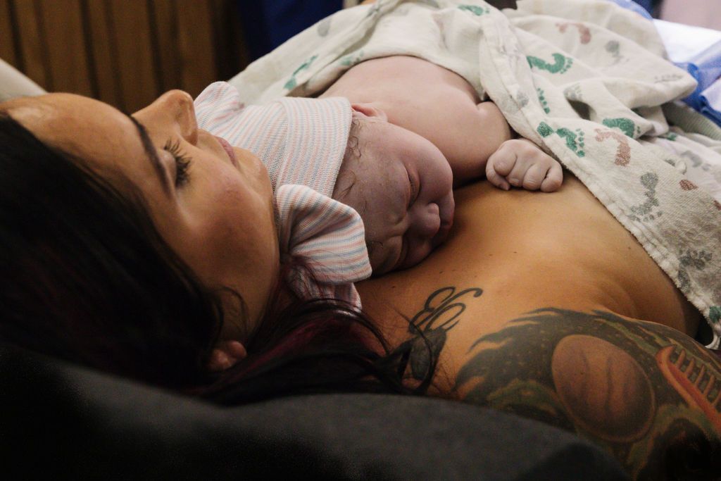New mother lying in hospital bed with tattooed arm holding her newborn baby on her chest after birth.