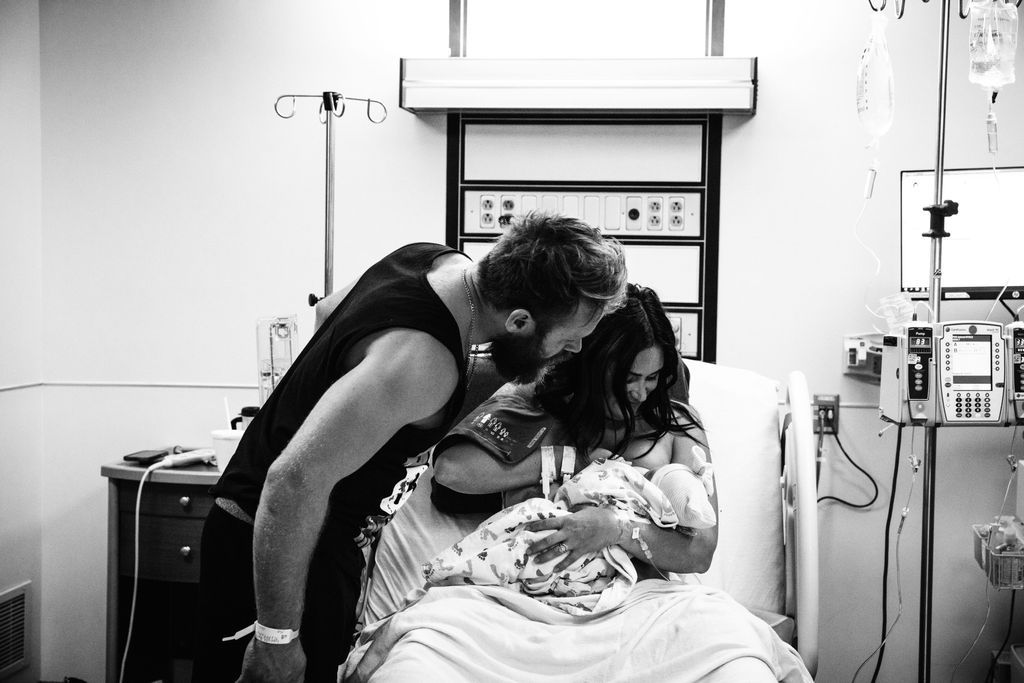 Black and white photo of new parents sharing a tender kiss over their newborn baby in a hospital room.