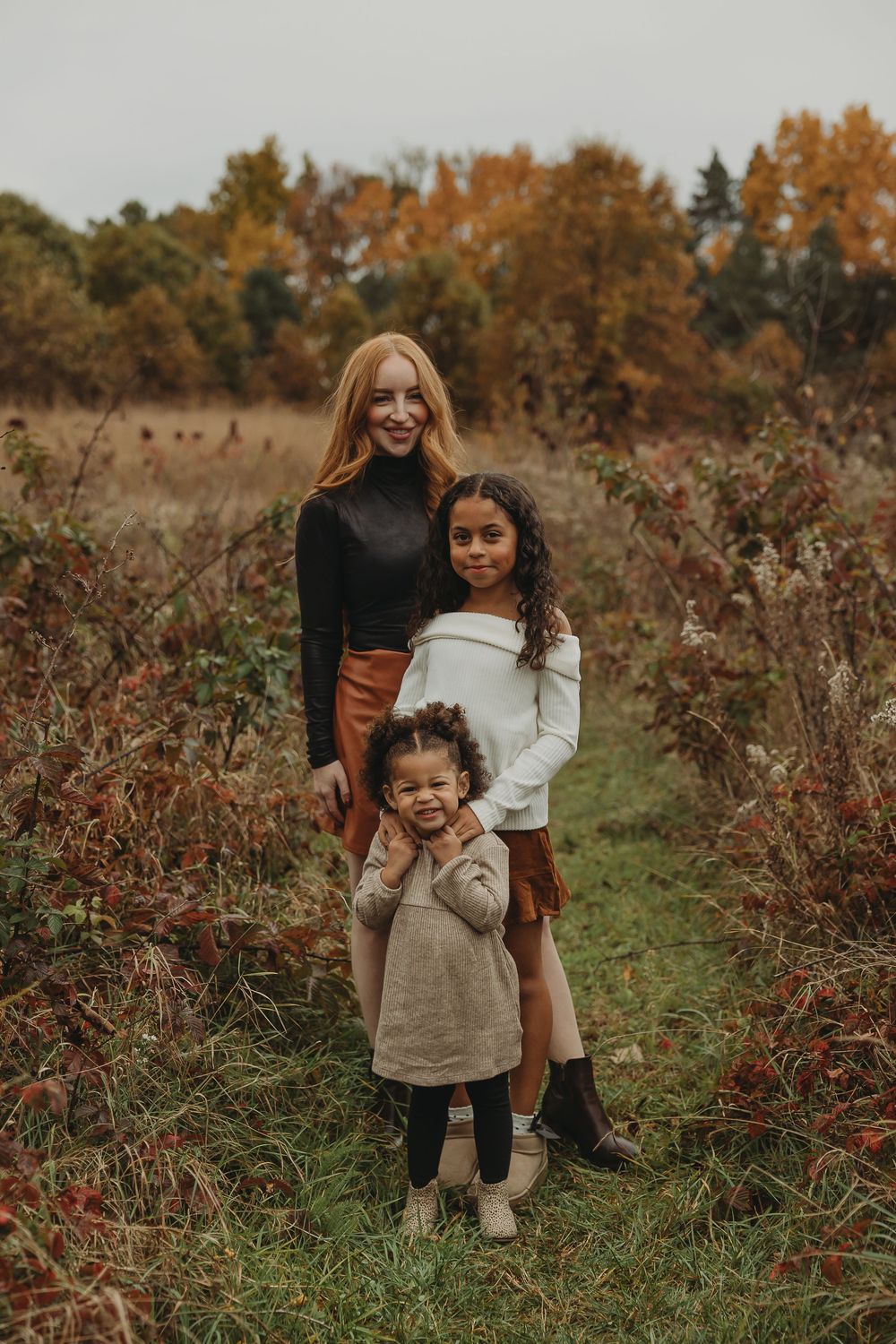 Three people pose together outdoors in a field during autumn with colorful fall foliage in the background.