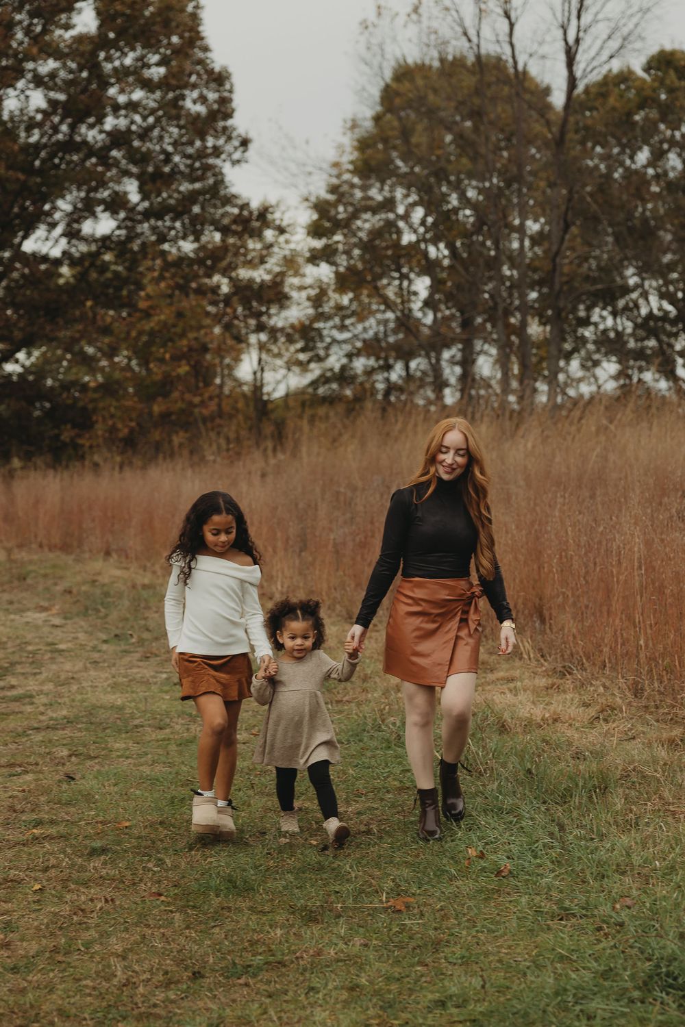 Three people walking together through a fall field with tall brown grass and trees in the background.