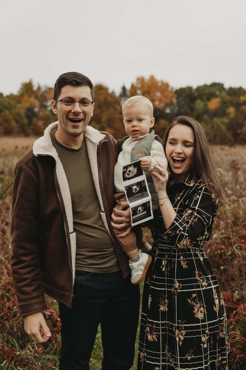 A family shares joyful moments in an autumn field while holding ultrasound photos during a pregnancy announcement photo shoot.