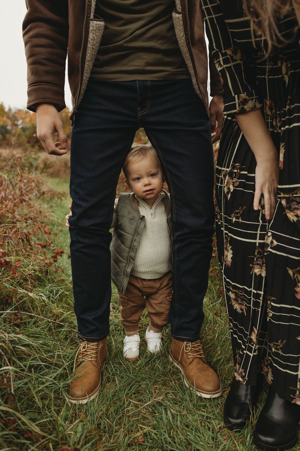 Small child peeks through parent's legs while standing in autumn field wearing neutral colored clothing.
