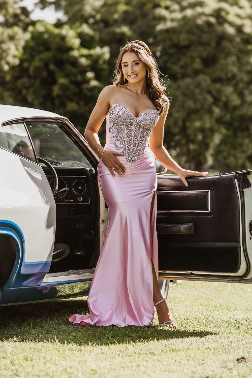 Formal/Graduation session young woman in elegant strapless pink and silver gown posing for photos beside a blue car outdoors in Caboolture.